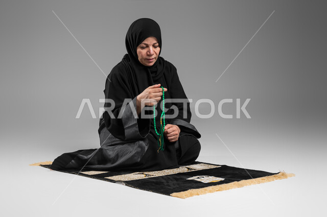 الإجتهاد في العباده والتقرب الى الله في شهر رمضان, مفهوم التسبيح والتهليل، بورتريه لسيدة عربية سعودية خليجية كبيرة بالسن ترتدي الحجاب والعباءة السوداء تجلس على سجادة الصلاة تمسك بيدها السبحه, الإكثار من ذكر الله تعالى من خلال الإستغفار والدعاء, خلفيه رمادية