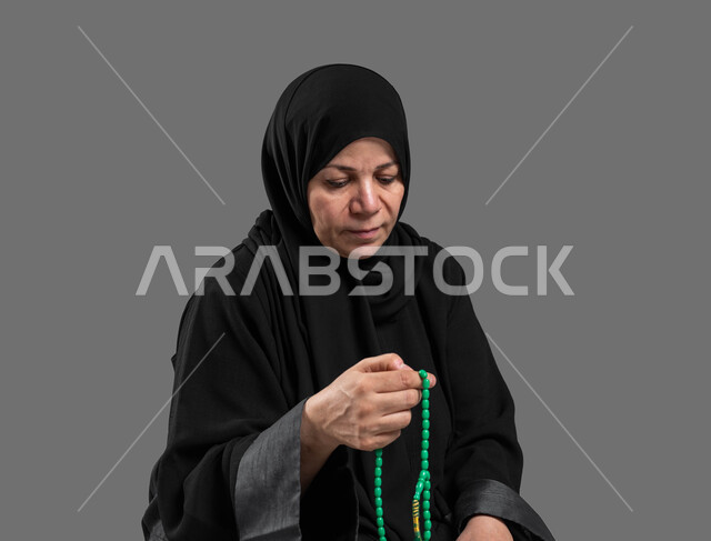 مفهوم التسبيح والتهليل، بورتريه مقرب لإمرأة عربية سعودية خليجية كبيرة بالسن ترتدي الحجاب والعباءة السوداء تمسك بيدها السبحه, الإكثار من ذكر الله تعالى من خلال الإستغفار والدعاء, الإجتهاد في العباده والتقرب الى الله في شهر رمضان, إسلاميات وعبادة, خلفيه رمادية