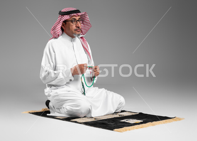 الإجتهاد في العباده والتقرب الى الله في شهر رمضان المبارك, مفهوم التسبيح والتهليل، بورتريه لرجل عربي سعودي خليجي كبير بالسن يرتدي الشماغ والثوب التقليدي يجلس على سجادة الصلاة يمسك بيده السبحه, الإكثار من ذكر الله تعالى من خلال الإستغفار والدعاء, خلفيه رمادية