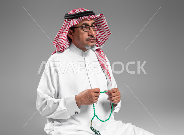 مفهوم التسبيح والتهليل، بورتريه مقرب لرجل عربي سعودي خليجي كبير بالسن يرتدي الشماغ والثوب التقليدي يمسك بيده السبحه, الإكثار من ذكر الله تعالى من خلال الإستغفار والدعاء, الإجتهاد في العباده والتقرب الى الله في شهر رمضان المبارك, إسلاميات وعبادة, خلفيه رمادية