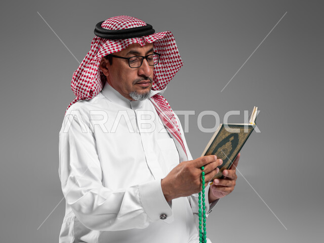 الإكثار من ذكر الله تعالى والإجتهاد في العباده, قراءة وتلاوة القرآن الكريم، بورتريه مقرب لرجل عربي سعودي خليجي كبير بالسن يرتدي الشماغ والثوب التقليدي ونظارة طبية يحمل بيده المصحف الشريف والسبحة, التقرب الى الله في شهر رمضان المبارك, خلفية رمادية