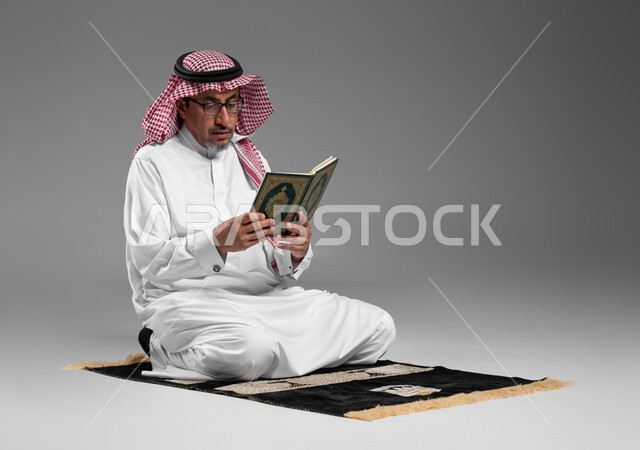 الشعور بالخشوع والطمأنينه, تلاوة وقراءة القرآن الكريم، بورتريه لرجل عربي سعودي خليجي يرتدي الشماغ والثوب التقليدي ونظارة طبية يجلس على سجادة الصلاة يحمل بيده المصحف الشريف, الإكثار من ذكر الله تعالى والإجتهاد في العباده, التقرب الى الله في شهر رمضان, خلفية رمادية