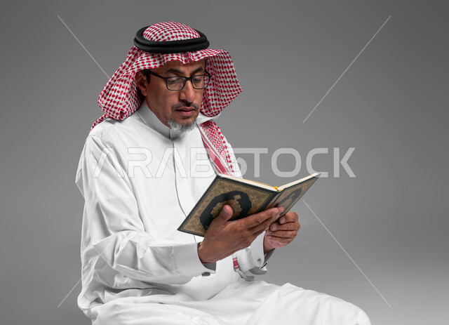 تلاوة وقراءة القرآن الكريم، بورتريه مقرب لرجل عربي سعودي خليجي كبير بالسن يرتدي الشماغ والثوب التقليدي ونظارة طبية يحمل بيده المصحف الشريف, الشعور بالخشوع والطمأنينه, الإكثار من ذكر الله تعالى والإجتهاد في العباده, التقرب الى الله في شهر رمضان, خلفية رمادية