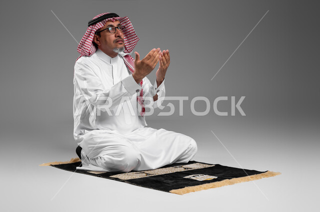 أداء فروض الصلاة على وقتها, التقرب الى الله من خلال الإجتهاد في العبادة والحفاظ على الصلاة، بورتريه لرجل عربي سعودي خليجي كبير بالسن يرتدي الشماغ والثوب التقليدي ونظارة طبية يجلس على سجادة الصلاة يرفع يديه ويدعو الله، إيماءات تدل على التوسل والتضرع، خلفية رمادية