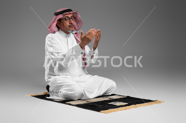 Commitment to praying on time, getting closer to God through diligence in worship and maintaining prayer, portrait of an elderly Saudi Arabian Gulf man wearing a shemagh, traditional dress, and medical glasses, sitting on the prayer rug, raising his hands and calling upon God, gestures indicating pleading and supplication, gray background.