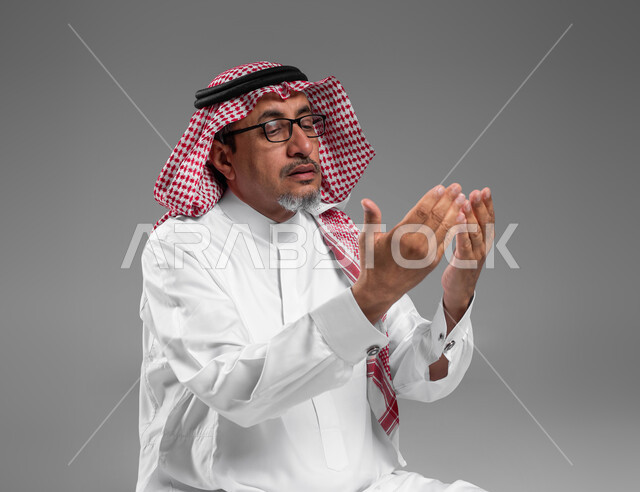 الإكثار من الدعاء في شهر رمضان المبارك، التقرب الى الله من خلال الإجتهاد في العبادة والحفاظ على الصلاة، بورتريه مقرب لرجل عربي سعودي خليجي كبير بالسن يرتدي الشماغ والثوب التقليدي ونظارة طبية يرفع يديه ويدعو الله بإيماءات تدل على التوسل والتضرع، خلفية رمادية