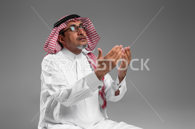 الإكثار من الدعاء في شهر رمضان المبارك، التقرب الى الله من خلال الإجتهاد في العبادة والحفاظ على الصلاة، بورتريه مقرب لرجل عربي سعودي خليجي كبير بالسن يرتدي الشماغ والثوب التقليدي ونظارة طبية يرفع يديه ويدعو الله بإيماءات تدل على التوسل والتضرع، خلفية رمادية