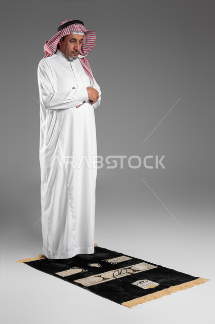 أذكار وأدعية وفرائض، الركوع والسجود والخشوع لله، بورتريه لرجل كبير السن عربي سعودي خليجي يرتدي الشماغ والثوب التقليدي يقف على السجادة و يؤدي فريضة الصلاة ، آداء صلاة التراويح ، الدعاء والتضرع الى الله، خلفية رمادية .