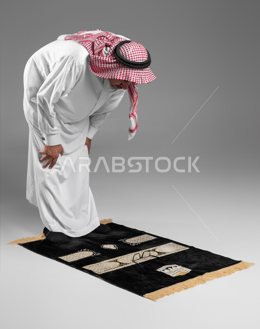 الركوع والسجود والخشوع لله، بورتريه لرجل كبير السن عربي سعودي خليجي يرتدي الشماغ والثوب التقليدي يقف على السجادة و يؤدي فريضة الصلاة ، آداء صلاة التراويح ، الدعاء والتضرع الى الله، أذكار وأدعية وفرائض، خلفية رمادية .