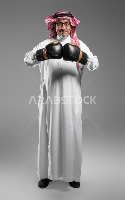 إتقان فنون الملاكمة، زيادة قدرة العضلات على التحمل، بورتريه لرجل كبير السن عربي خليجي سعودي مبتسم يرتدي القفازات ينظر إلى الكاميرا بإيماءات القوة، ممارسة رياضة الدفاع عن النفس، تعزيز صحة العظام والمفاصل، صورة الجسم بالطول الكامل، خلفية رمادية .
