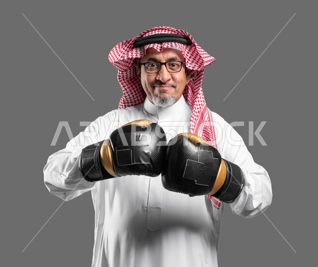 أنشطة بدنية للمحافظة على لياقة الجسم، بورتريه مقرب لرجل كبير السن عربي خليجي سعودي مبتسم يرتدي القفازات ينظر إلى الكاميرا بإيماءات القوة والثقة بالنفس، ممارسة رياضة الدفاع عن النفس، التدرب على الحركات القتالية، تعزيز صحة العظام والمفاصل، خلفية رمادية .