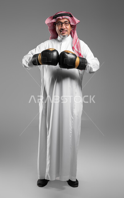 Resistance and increasing muscle endurance, a portrait of an old, smiling Saudi Gulf Arab man wearing gloves looking at the camera with force gestures, practicing self-defense, practicing combat movements, enhancing bone and joint health, full-length body portrait, gray background
