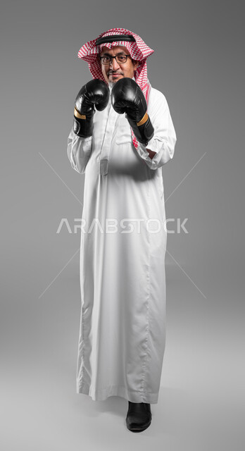 دعم الثقة بالنفس وتقليل الشعور بالتوتر، زيادة قدرة العضلات على التحمل، بورتريه مقرب لرجل كبير السن عربي خليجي سعودي مبتسم يرتدي القفازات ينظر إلى الكاميرا يتعلم فنون الملاكمة، تعزيز صحة العظام والمفاصل، صورة الجسم بالطول الكامل، خلفية رمادية .