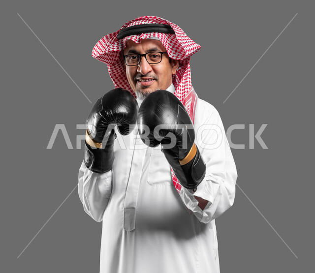 تقليل الشعور بالتوتر ودعم الثقة بالنفس، زيادة قدرة العضلات على التحمل، بورتريه مقرب لرجل كبير السن عربي خليجي سعودي مبتسم يرتدي القفازات ينظر إلى الكاميرا يتعلم فنون الملاكمة،  المحافظة على اللياقة البدنية، تعزيز صحة العظام والمفاصل، خلفية رمادية .