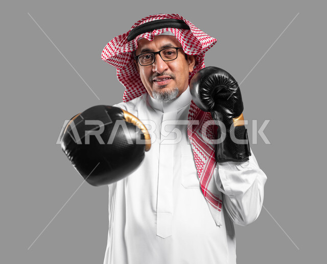المحافظة على اللياقة البدنية، زيادة قدرة العضلات على التحمل، بورتريه لرجل كبير السن عربي خليجي سعودي مبتسم يرتدي القفازات ينظر إلى الكاميرا يتعلم فنون الملاكمة، التدرب  على الحركات القتالية ، تعزيز صحة العظام والمفاصل، خلفية رمادية .
