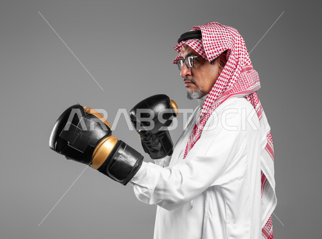 الاستعداد للتصدي والمقاومة، زيادة قدرة العضلات على التحمل، بورتريه مقرب لرجل كبير السن عربي خليجي سعودي يرتدي القفازات يتعلم فنون الملاكمة، ممارسة رياضة الدفاع عن النفس، التدرب على الحركات القتالية، المحافظة على اللياقة البدنية، خلفية رمادية .