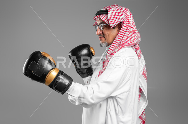 زيادة قدرة العضلات على التحمل، بورتريه مقرب لرجل كبير السن عربي خليجي سعودي مبتسم يرتدي القفازات يتعلم فنون الملاكمة، ممارسة رياضة الدفاع عن النفس، التدرب  على الحركات القتالية ، المحافظة على اللياقة البدنية، تعزيز صحة العظام والمفاصل، خلفية رمادية .
