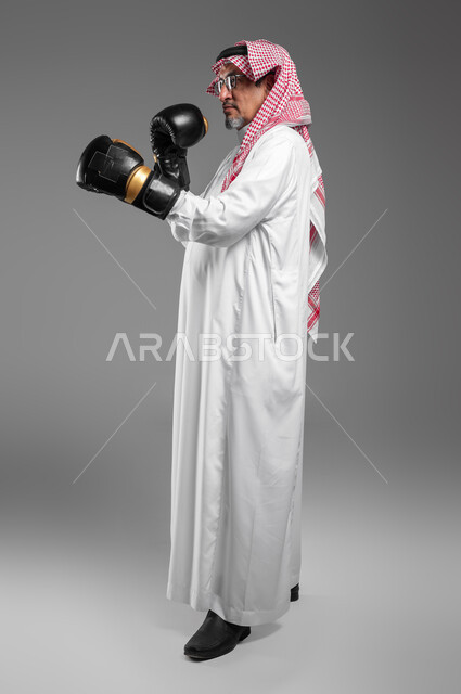 ممارسة رياضة الدفاع عن النفس، زيادة قدرة العضلات على التحمل، بورتريه لرجل كبير السن عربي خليجي سعودي يرتدي القفازات يتعلم فنون الملاكمة، التدرب  على الحركات القتالية ، المحافظة على اللياقة البدنية، تعزيز صحة العظام والمفاصل، صورة الجسم بالطول الكامل, خلفية رمادية .