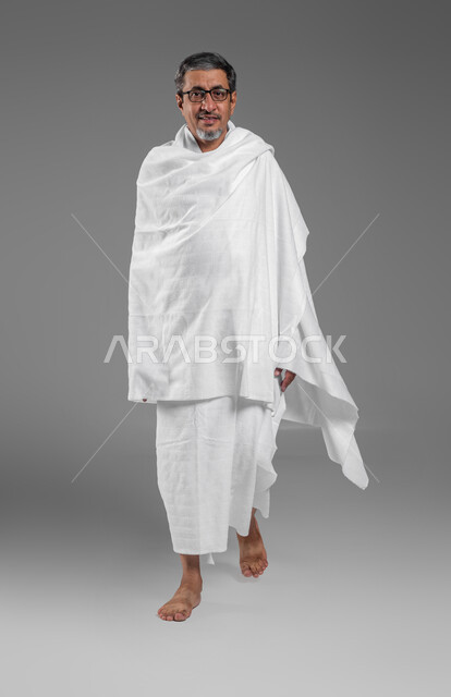 ايماءات المشي والذهاب الى مكان ما، الاستعداد لأداء مناسك الحج في شهر ذو الحجة، بورتريه لرجل عربي خليجي سعودي مبتسم يرتدي لباس الاحرام يقوم بزيارة بيت الله الحرام في مكة المكرمة، العبادة والتقرب الى الله، صورة الجسم بالطول الكامل، خلفية رمادية