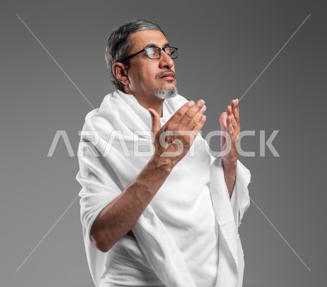 تلبية نداء الله لاداء فريضة الحج، المحافظة على الدعاء وطلب المغفرة من الله تعالى, فضل العمرة في شهر رمضان المبارك,  بورتريه مقرب لرجل عربي خليجي سعودي يرتدي ثياب الاحرام يدعو بخشوع وتضرع، اسلاميات وعبادة، خلفية رمادية.