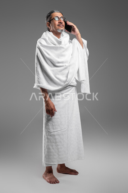 Answering calls via mobile phone, portrait of an elderly Gulf Arab Saudi man, smiling, wearing Ihram clothes preparing to visit the Holy House of God, getting closer to God by performing the duties of Hajj and Umrah, gestures of happiness in communicating with family and friends, full body portrait, gray background