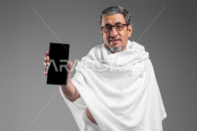 الحجز لزيارة بيت الله الحرام اونلاين عبر الانترنت، تطبيقات وبرامج الهاتف المحمول لارشادات الحج والعمرة, بورتريه مقرب لرجل مسن عربي سعودي خليجي مبتسم يرتدي ثياب الاحرام يقوم بعرض شاشة جوال سوداء فارغة، الدليل الالكتروني للحاج والمعتمر، خلفية رماديه