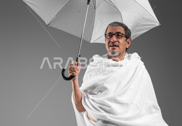 زيارة المشاعر المقدسة في مكة المكرمة، بورتريه مقرب لرجل مسن عربي خليجي سعودي  مبتسم يرتدي ثياب الاحرام يحمل بيده مظلة شمسية للحماية والتظلل، تعابير السرور بالاستعداد لأداء فريضة الحج، العبادة والتقرب الى الله، السفر للعمره في شهر رمضان المبارك, خلفية رمادية