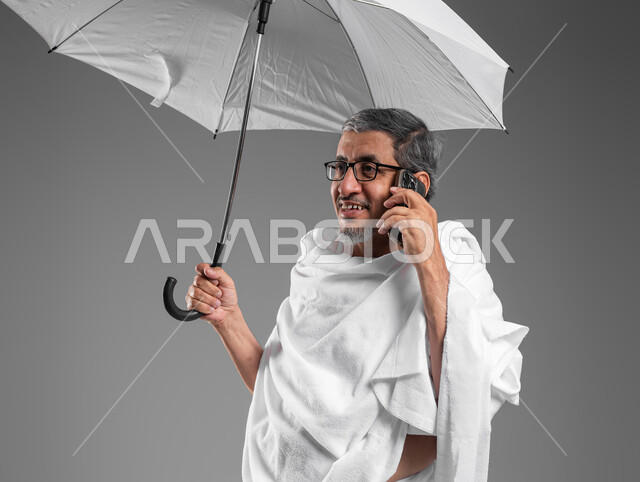 ايماءات تدل على السعادة بالتواصل مع العائلة والأصدقاء، بورتريه مقرب لرجل مسن خليجي عربي سعودي مبتسم يرتدي ثياب الاحرام يحمل بيده مظلة شمسية بيضاء يجري مكالمة هاتفية باستخدام الهاتف المحمول، التقرب الى الله من خلال أداء فريضة الحج والعمرة، خلفية رمادية
