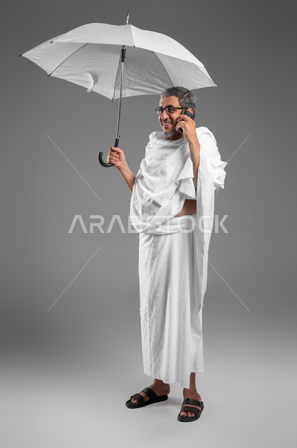 Answering calls via mobile phone, expressions of pleasure in communicating with family and friends, portrait of an elderly Gulf Arab Saudi man smiling, wearing Ihram clothes and holding a white umbrella in his hand, getting closer to God by performing the Hajj and Umrah duties, full body portrait, gray background