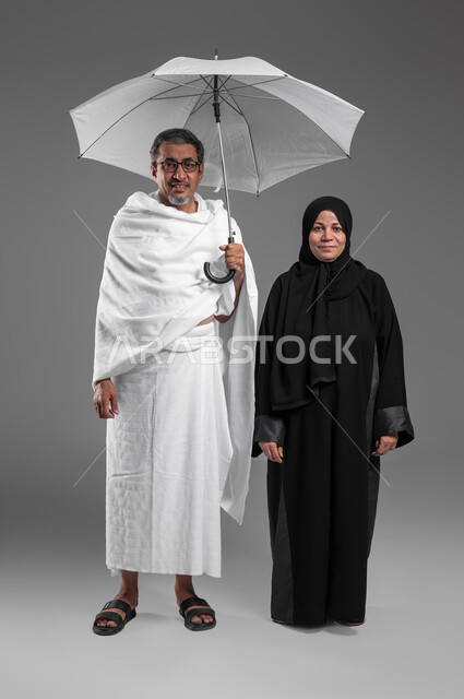 استخدام المظلة الشمسية للحماية والتظلل، تعابير السرور بالاستعداد لأداء فريضة الحج، بورتريه لزوجين عربيين خليجيين سعوديين كبيرين بالسن يرتديان ثياب الاحرام يقفان بإستقامة، التقرب الى الله تعالى، أداء العمرة في شهر رمضان المبارك، صورة الجسم بالكامل، خلفية رمادية