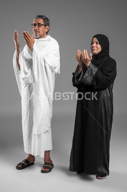 فضل العمرة في شهر رمضان المبارك, المحافظة على الدعاء وطلب المغفرة من الله تعالى, تلبية نداء الله لاداء فريضة الحج، بورتريه لزوجين عربيين خليجيين سعوديين كبيرين بالسن يرتديان ثياب الإحرام يدعوان بخشوع وتضرع، اسلاميات وعبادة، صورة الجسم بالكامل، خلفية رمادية.