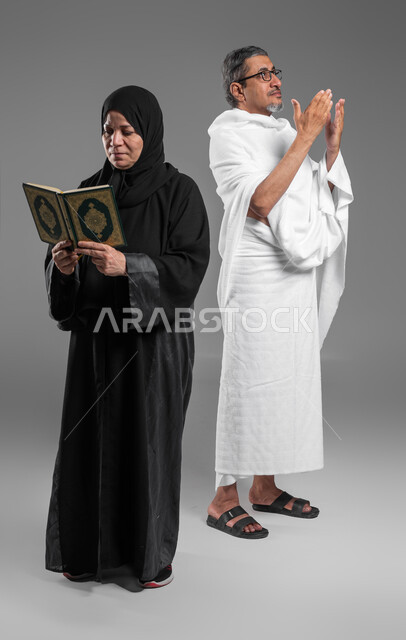 قراءة القرآن من المصحف الشريف, اداء مناسك الحج والعمرة، المحافظة على الدعاء وطلب المغفرة من الله تعالى, بورتريه لرجل وإمرأة عربيين خليجيين سعوديين كبيريين بالسن يرتديان ثياب الاحرام يقومان بالتقرب الى الله بالاعمال الصالحة، صورة الجسم بالكامل، خلفية رمادية