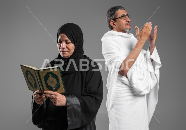 أداء مناسك الحج والعمرة، قراءة القرآن من المصحف الشريف, المحافظة على الدعاء وطلب المغفرة من الله تعالى, بورتريه مقرب لرجل وإمرأة عربيين خليجيين سعوديين كبيريين بالسن يرتديان ثياب الاحرام يقومان بالتقرب الى الله بالاعمال الصالحة، اسلاميات وعبادة، خلفية رمادية