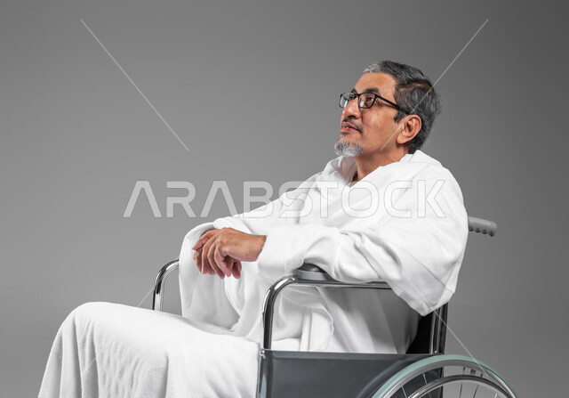 ايماءات السرور والانشراح بأداء مناسك الحج والعمرة، الركن الخامس من اركان الاسلام، بورتريه مقرب لرجل كبير بالسن عربي سعودي خليجي مبتسم مقعد يرتدي ثياب الاحرام يجلس على كرسي متحرك، العبادة والتقرب الى الله، خلفية رمادية