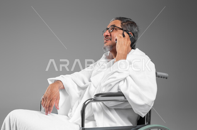 الرد على الاتصالات عبر الهاتف المحمول، ايماءات تدل على السعادة بالتواصل مع العائلة والأصدقاء، بورتريه مقرب لرجل مسن خليجي عربي سعودي مقعد يرتدي ثياب الاحرام يجلس على كرسي متحرك يستعد لاداء فريضة الحج والعمرة، التقرب الى الله من خلال أداء الفرائض، خلفية رمادية