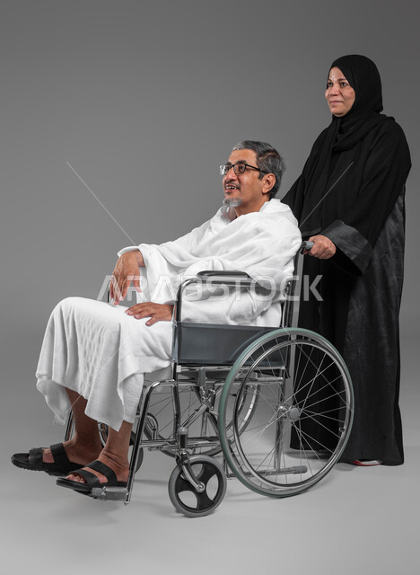 مساعدة كبار السن وذوي الاحتياجات الخاصه, السفر لاداء فريضة الحج في شهر ذو الحجه، بورتريه لرجل وامرأة عربيين خليجيين سعوديين يرتديان ملابس الاحرام يستعدان للعمره في شهر رمضان المبارك، مفهوم حسن المعاشره في الحياه الزوجيه, صورة الجسم بالكامل، خلفيه رماديه