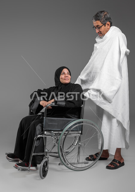 مفهوم حسن المعاشره  في الحياه الزوجيه, بورتريه لرجل وامرأة عربيين خليجيين سعوديين يرتديان ملابس الاحرام يستعدان لاداء فريضة الحج في شهر ذو الحجه, مساعدة ذوي الاحتياجات الخاصه وكبار السن, السفر للعمره في شهر رمضان المبارك, صورة الجسم بالكامل، خلفيه رماديه