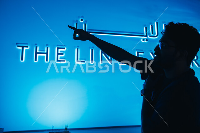 بناء مستقبل حضاري متطور في المملكة، رجل عربي خليجي سعودي يقف أمام شعار معرض "ذا لاين" في الرياض ويشير إلى شيء ما، التخطيط المعماري لقطاع العقارات، الابتكارات المستقبلية في البنية التحتية للمدن الذكية، رؤية سعودية مستقبلية 2030