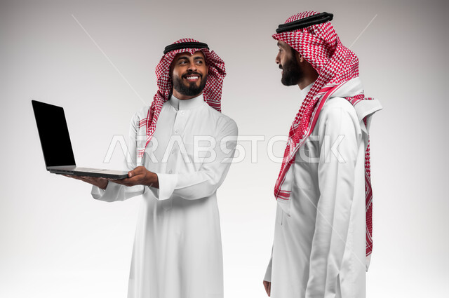 التعاون بين الموظفين لتحقيق أهداف الشركة، متابعة مشاريع الأعمال على الحاسب المحمول، بورتريه مقرب لرجلي أعمال سعوديين خليجيين عربيين يرتديان الشماغ والثوب التقليدي يقومان بإنجاز مهام العمل عبر جهاز الحاسوب، دمج العمل مع التقنية والتكنولوجيا، خلفية بيضاء