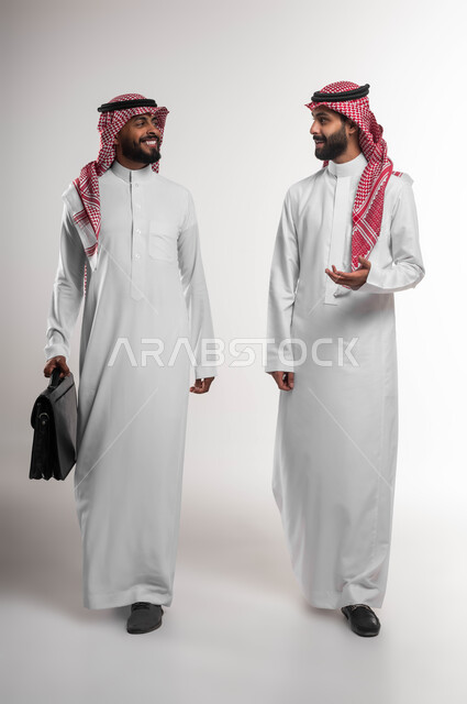 التعاون بين الموظفين لتحقيق أهداف الشركة، الاهتمام بالأناقة والمظهر الخارجي، الذهاب إلى دوام العمل بنشاط, بورتريه لرجلي أعمال سعوديين خليجيين عربيين يرتديان الشماغ والثوب التقليدي يتحدثان, صورة الجسم بالكامل, خلفية بيضاء