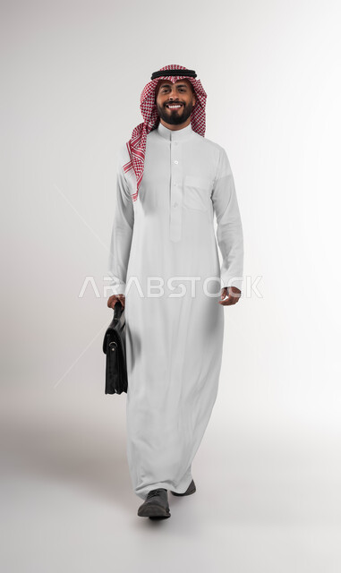 الشعور بالسرور لتحقيق أهداف الشركة, الذهاب إلى دوام العمل بنشاط, بورتريه لرجل عربي سعودي خليجي مبتسم يرتدي الشماغ والثوب التقليدي ينظر إلى الكاميرا يحمل حقيبة يد, الاهتمام بالأناقة والمظهر الخارجي، صورة الجسم بالكامل, خلفية بيضاء