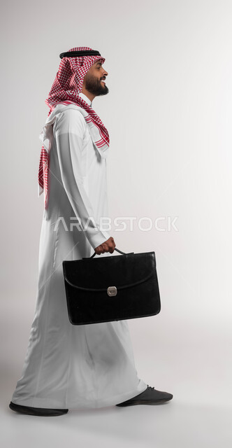 الذهاب إلى دوام العمل بنشاط, الشعور بالسرور لتحقيق أهداف الشركة, بورتريه من الجانب لرجل أعمال عربي سعودي خليجي مبتسم يرتدي الشماغ والثوب التقليدي يحمل حقيبة يد, الاهتمام بالأناقة والمظهر الخارجي، صورة الجسم بالكامل, خلفية بيضاء