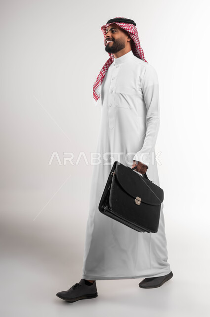 الذهاب إلى دوام العمل بنشاط, الشعور بالسرور لتحقيق أهداف الشركة, بورتريه من الجانب لرجل أعمال عربي سعودي خليجي مبتسم يرتدي الشماغ والثوب التقليدي يحمل حقيبة يد, الاهتمام بالأناقة والمظهر الخارجي، صورة الجسم بالكامل, خلفية بيضاء