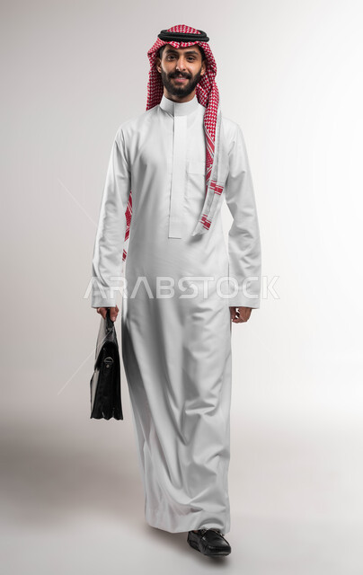 الشعور بالسرور لتحقيق أهداف الشركة, الذهاب إلى دوام العمل بنشاط, بورتريه لرجل عربي سعودي خليجي مبتسم يرتدي الشماغ والثوب التقليدي ينظر إلى الكاميرا يحمل حقيبة يد, الاهتمام بالأناقة والمظهر الخارجي، صورة الجسم بالكامل, خلفية بيضاء