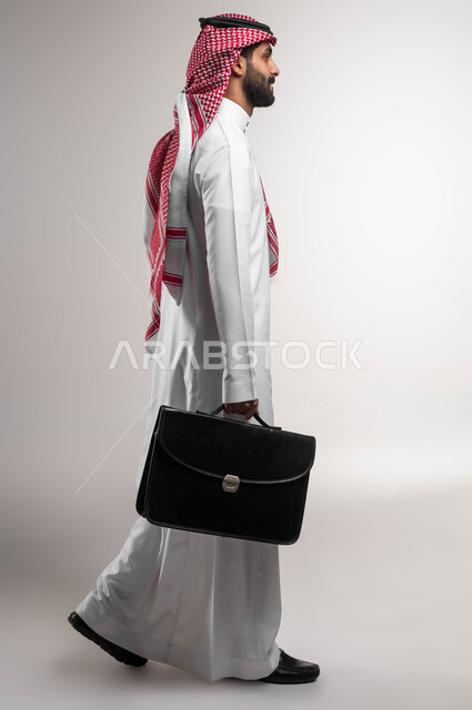 الاهتمام بالأناقة والمظهر الخارجي، الذهاب إلى دوام العمل بنشاط, الشعور بالسرور لتحقيق أهداف الشركة, بورتريه من الجانب لرجل أعمال عربي سعودي خليجي مبتسم يرتدي الشماغ والثوب التقليدي يحمل حقيبة يد, صورة الجسم بالكامل, خلفية بيضاء