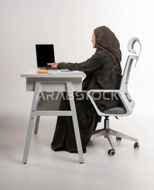 التفكير في استراتيجيات وأهداف العمل، إدارة شؤون الأعمال وتنظيمها, النقر والتفاعل مع لوحة المفاتيح, بورتريه من الجانب لموظفة عربية سعودية خليجية محجبة ترتدي العباءة تقوم بالعمل على جهاز اللابتوب المحمول بإيماءات التركيز والتمعن, خلفية بيضاء.