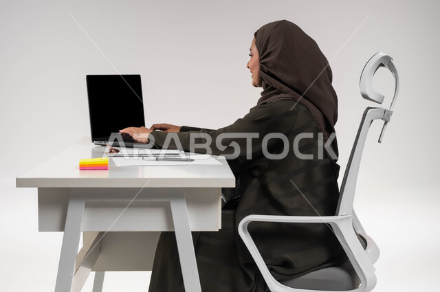 إدارة شؤون الأعمال وتنظيمها, النقر والتفاعل مع لوحة المفاتيح, التفكير في استراتيجيات وأهداف العمل، بورتريه مقرب من الجانب لموظفة عربية سعودية خليجية محجبة ترتدي العباءة تقوم بالعمل على جهاز اللابتوب المحمول بإيماءات التركيز والتمعن, خلفية بيضاء .