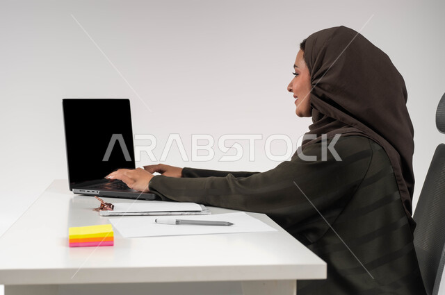 إدارة شؤون الأعمال وتنظيمها, النقر والتفاعل مع لوحة المفاتيح, التفكير في استراتيجيات وأهداف العمل، بورتريه مقرب من الجانب لموظفة عربية سعودية خليجية محجبة ترتدي العباءة تقوم بالعمل على جهاز اللابتوب المحمول بإيماءات التركيز والتمعن, خلفية بيضاء .