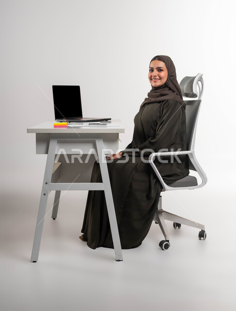 دمج التقنية والتكنولوجيا مع العمل، مهن ووظائف مكتبية نسائية، بورتريه لموظفة عربية سعودية خليجية محجبة مبتسمة تجلس أمام المكتب وتنظر إلى الكاميرا بإيماءات الثقة بالنفس, الاهتمام بالأناقة والمظهر الخارجي، خلفية بيضاء.