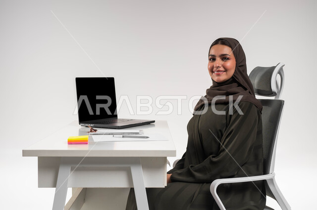مهن ووظائف مكتبية نسائية، دمج التقنية والتكنولوجيا مع العمل، بورتريه مقرب لموظفة عربية سعودية خليجية محجبة مبتسمة تجلس أمام المكتب وتنظر إلى الكاميرا بإيماءات الثقة بالنفس, الاهتمام بالأناقة والمظهر الخارجي، خلفية بيضاء.